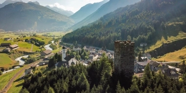 View of a picturesque valley with a river, medieval tower, and a church, surrounded by mountains and forests.