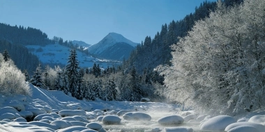 Snow-covered river with snow-capped rocks, surrounded by a winter forest and mountains in the background.