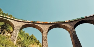 Brücke mit Viadukt-Bögen und Zug darauf, vor blauem Himmel.