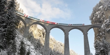 A red train crosses an impressive stone viaduct in the snowy mountains.