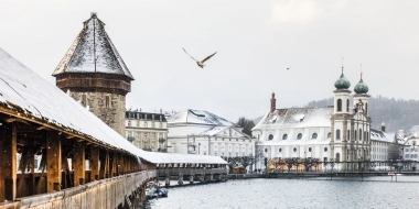 Wooden bridge over a river with a tower and snow-covered buildings in the background.