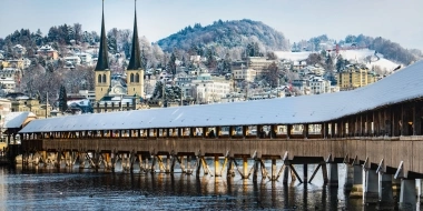 A snowy wooden bridge crosses a river in a picturesque town with churches and hills in the background.