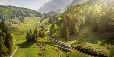 A train travels through a green, mountainous landscape with forests and meadows.