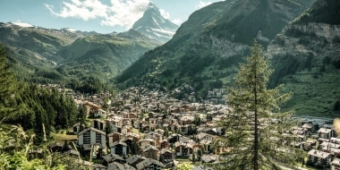 Blick auf ein alpines Tal mit einer Stadt, umgeben von grünen Bergen und einem markanten Berggipfel im Hintergrund.