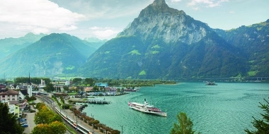 Paysage de montagne avec lac, train et bateau au premier plan.