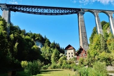 Eine große Eisenbahnbrücke überquert ein grünes Tal mit einem roten Haus darunter.