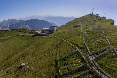 Une station de montagne sur une colline verte avec des sentiers de randonnée et des antennes, avec une chaîne de montagnes en arrière-plan.