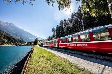 A red Bernina Express train travels along a blue lake in a mountain landscape in sunlight.