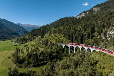 Eine rote Bahn überquert ein Viadukt in einer bergigen und bewaldeten Landschaft unter klarem, blauem Himmel.