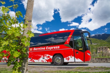 A red Bernina Express bus on a road in front of a mountain landscape under a blue sky.