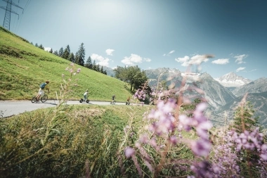 Eine Gruppe von Menschen fährt auf E-Scootern einen Bergpfad entlang, umgeben von blühenden Wiesen und Bergen im Hintergrund.