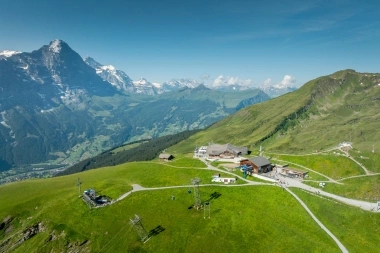 Vue paysagère d'une station de montagne alpine avec des prairies vertes et des montagnes enneigées en arrière-plan.