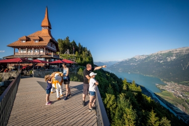 A family on a pier overlooking a lake and mountains, with a building with a red roof in the background.