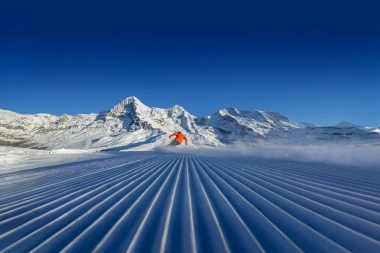 Skier in orange on freshly groomed slope in front of snow-covered mountains.