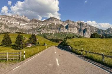 Route de campagne avec prairies vertes et montagnes majestueuses en arrière-plan sous un ciel bleu.