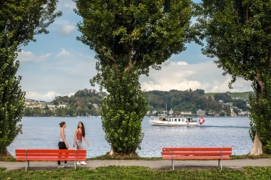 Two people walking along red benches, with trees and a ship on the lake in the background.