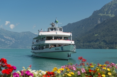 A ship on a lake surrounded by mountains; colorful flowers in the foreground.