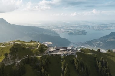 Aerial view of a mountain peak with a building, surrounded by green hills and a lake in the background.