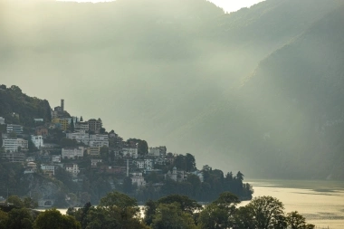 A city on a hill surrounded by mist, with a lake in the foreground and wooded mountains in the background.