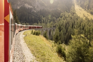 A red train is traveling on a viaduct through a mountainous forest landscape.