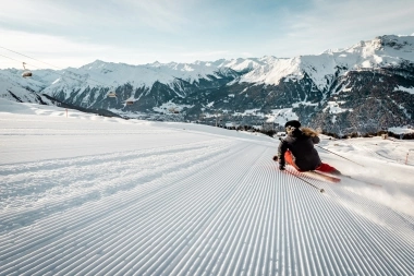 Una sciatrice su una pista battuta con montagne innevate sullo sfondo.