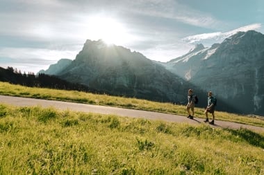 Two hikers on a trail with mountains in the background and sunshine.