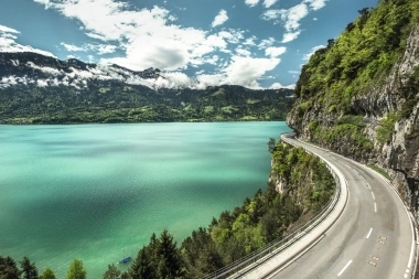 Eine kurvige Strasse an einem türkisfarbenen See, umgeben von Bergen und Wald.