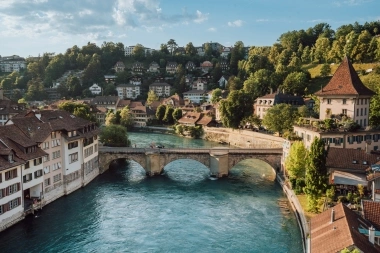 Eine hübsche Altstadt an einem Fluss mit einer steinernen Brücke und grünen Hügeln im Hintergrund.