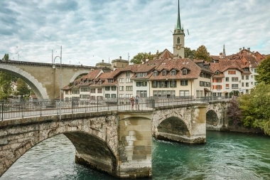 Un ponte di pietra attraversa un fiume, con edifici storici e un campanile sullo sfondo.