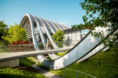 A modern, arched glass architecture with a walkway and trees in the foreground.