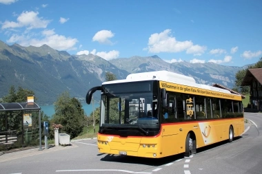 A yellow bus on a winding road with mountains and a lake in the background.