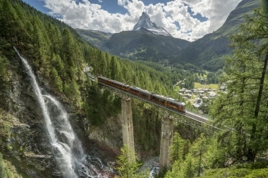 A train crosses a bridge with a waterfall in the foreground and a mountain in the background.