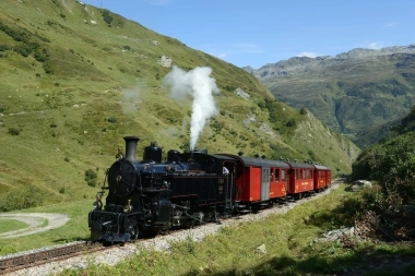 Eine schwarze Dampflokomotive zieht rote Personenzüge durch eine grüne Berglandschaft.