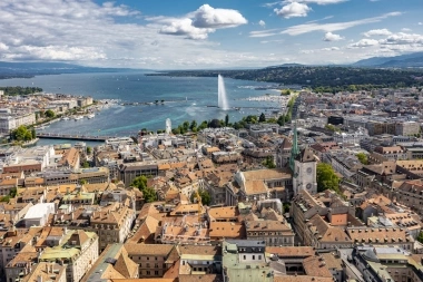 Luftaufnahme einer Stadt an einem See mit einem grossen Springbrunnen und umliegenden Hügeln.