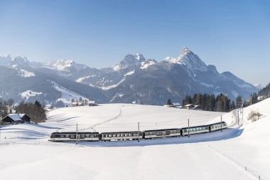 A winter mountain landscape with a train traveling through snowy fields.