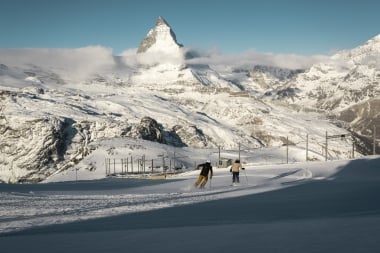 Zwei Skifahrer auf einer Piste mit dem verschneiten Matterhorn im Hintergrund.