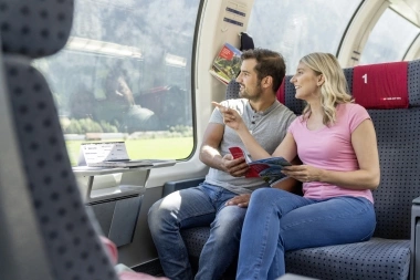 A couple sits on a train, looking out the window and holding travel guides in their hands.