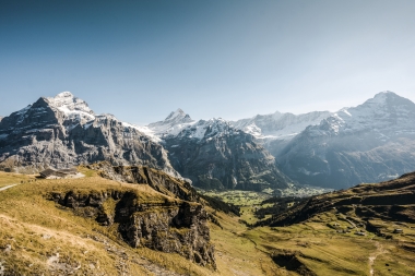 Eine weite Berglandschaft mit schneebedeckten Gipfeln und grünen Tälern unter klarem Himmel.