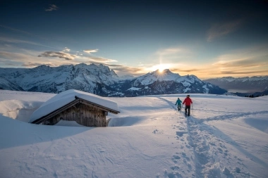 Zwei Personen wandern im Schnee bei Sonnenuntergang, Berge im Hintergrund.