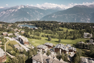 Aerial view of an alpine landscape with houses, a lake, and mountains in the background.