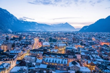 A winter cityscape with lit streets, surrounded by snow-covered mountains at dusk.