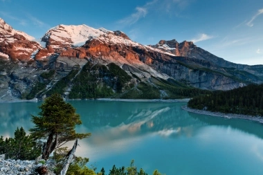 A tranquil mountain landscape with a turquoise lake and snow-covered peaks in the background at sunset.