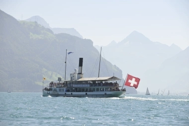 A steamboat with a Swiss flag sails on a lake surrounded by mountains.