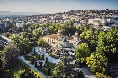 Aerial view of a historic building with a garden, surrounded by trees and a city in the background.