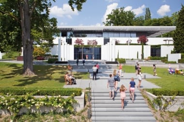 A modern white building facade with several columns, people in the forecourt, and a blooming garden landscape.