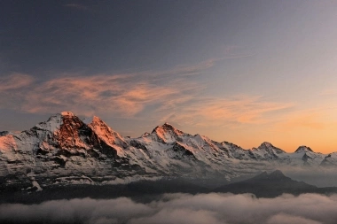 A mountain range at sunrise with snow-covered peaks and a slightly cloudy sky.