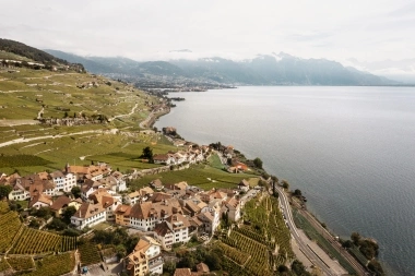 Aerial view of a village by the lake with terraced vineyards and mountains in the background.