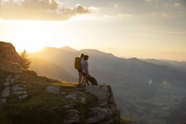 Two hikers stand on a mountain peak at sunset.