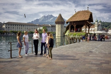 Eine Gruppe von Menschen steht auf einer Uferpromenade vor einer historischen Brücke mit Berg im Hintergrund.