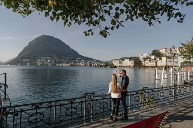 A couple stands by a railing by the lake, overlooking a city and a mountain in the background.